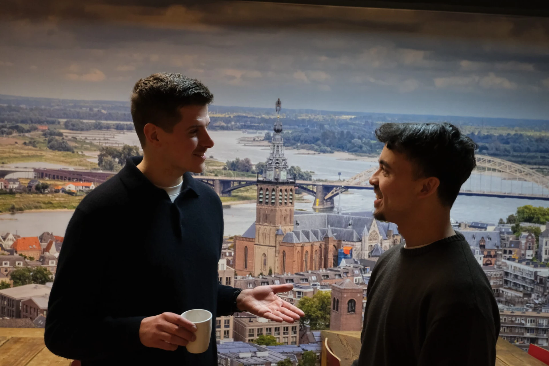 Twee mannen in gesprek voor een muurprint van Nijmegen met de Stevenskerk.