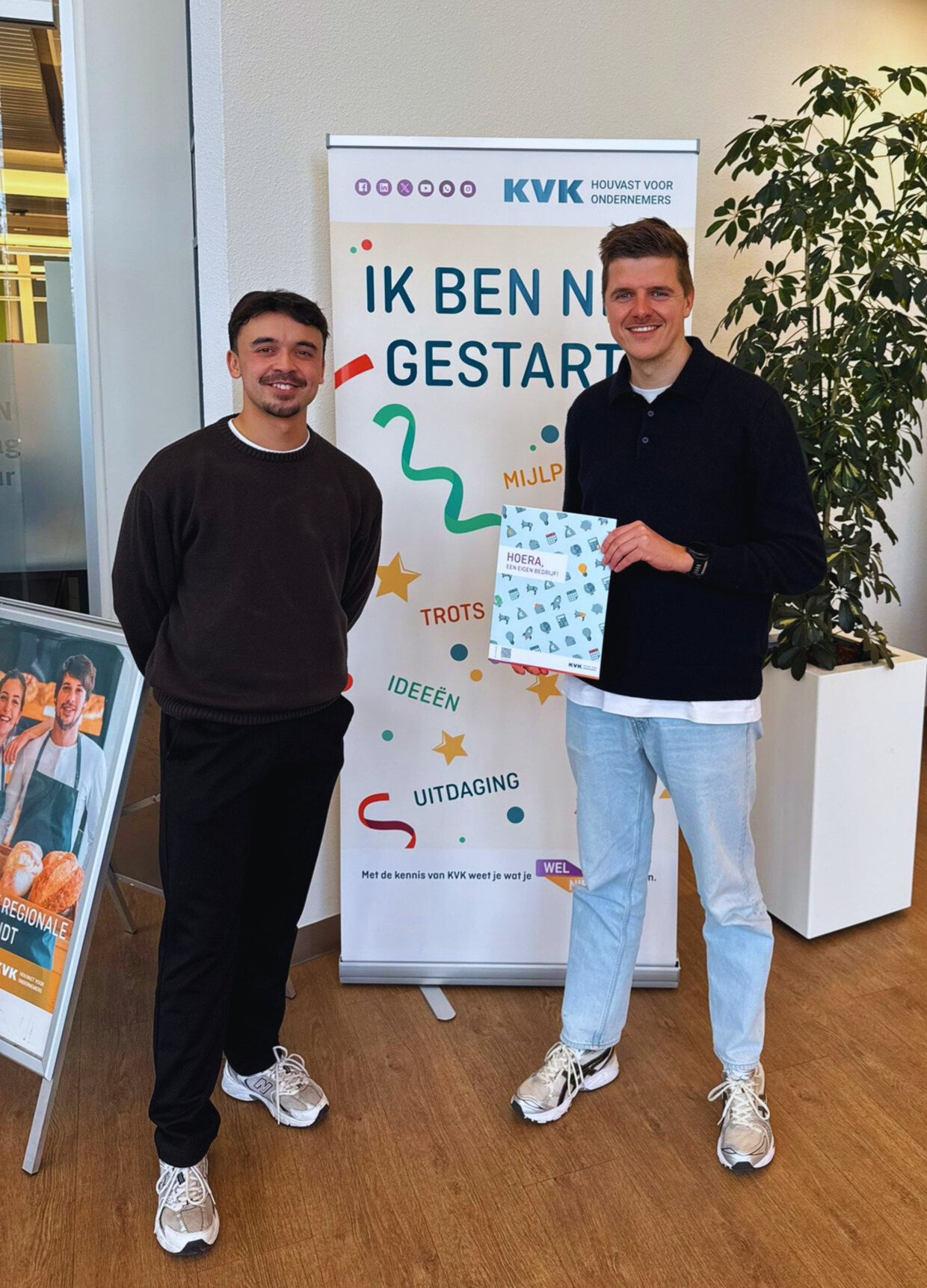 Twee glimlachende mannen voor een KVK-banner, van wie één een startersbrochure vasthoudt.
