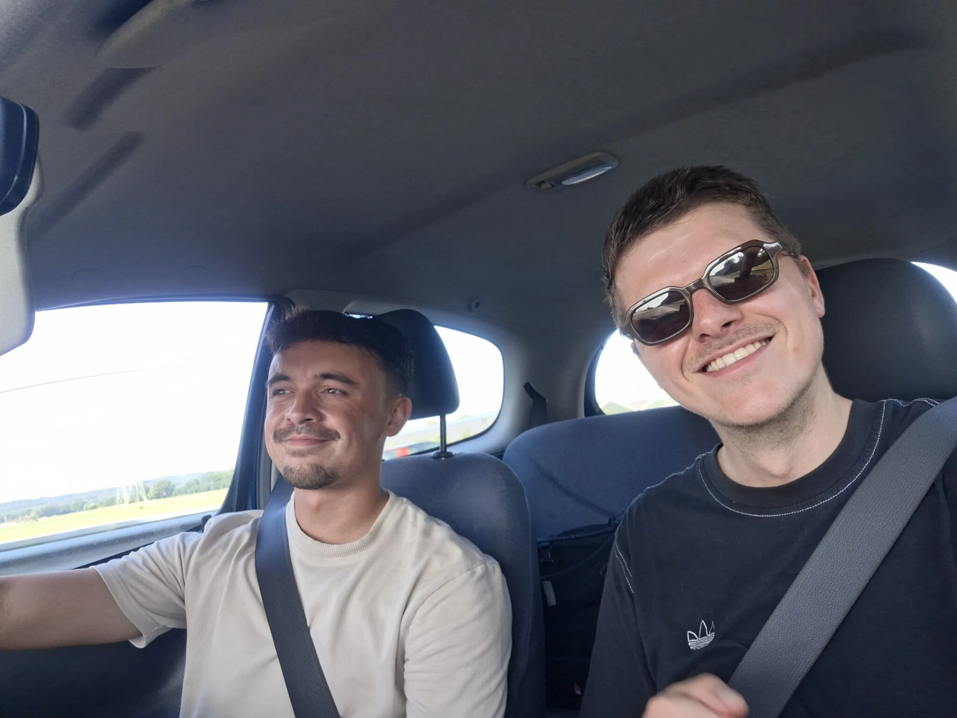 Twee mannen in een auto; de bestuurder kijkt naar de weg en de bijrijder met zonnebril lacht naar de camera.