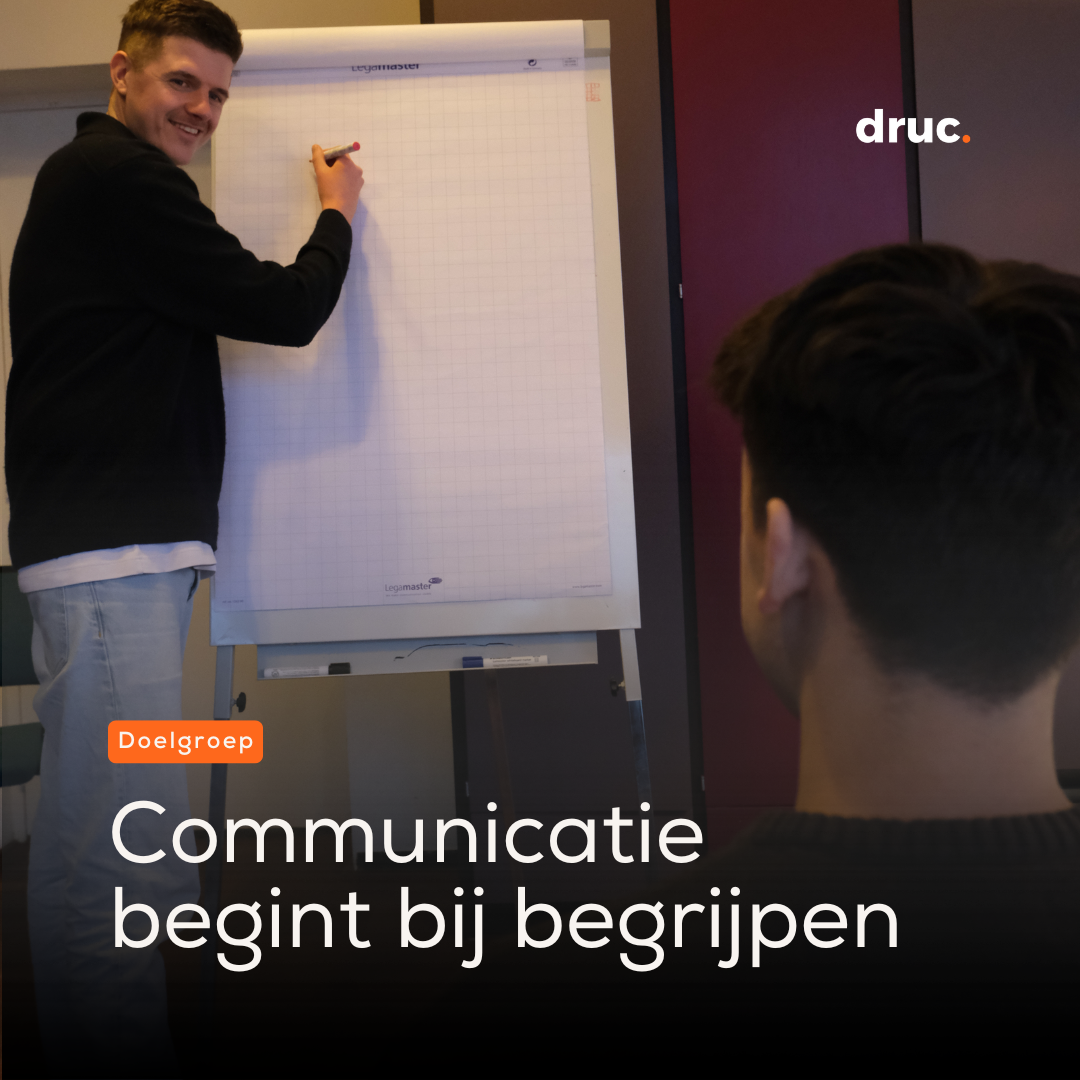 Een man staat met een stift bij een flipover en kijkt glimlachend naar een collega op de voorgrond.