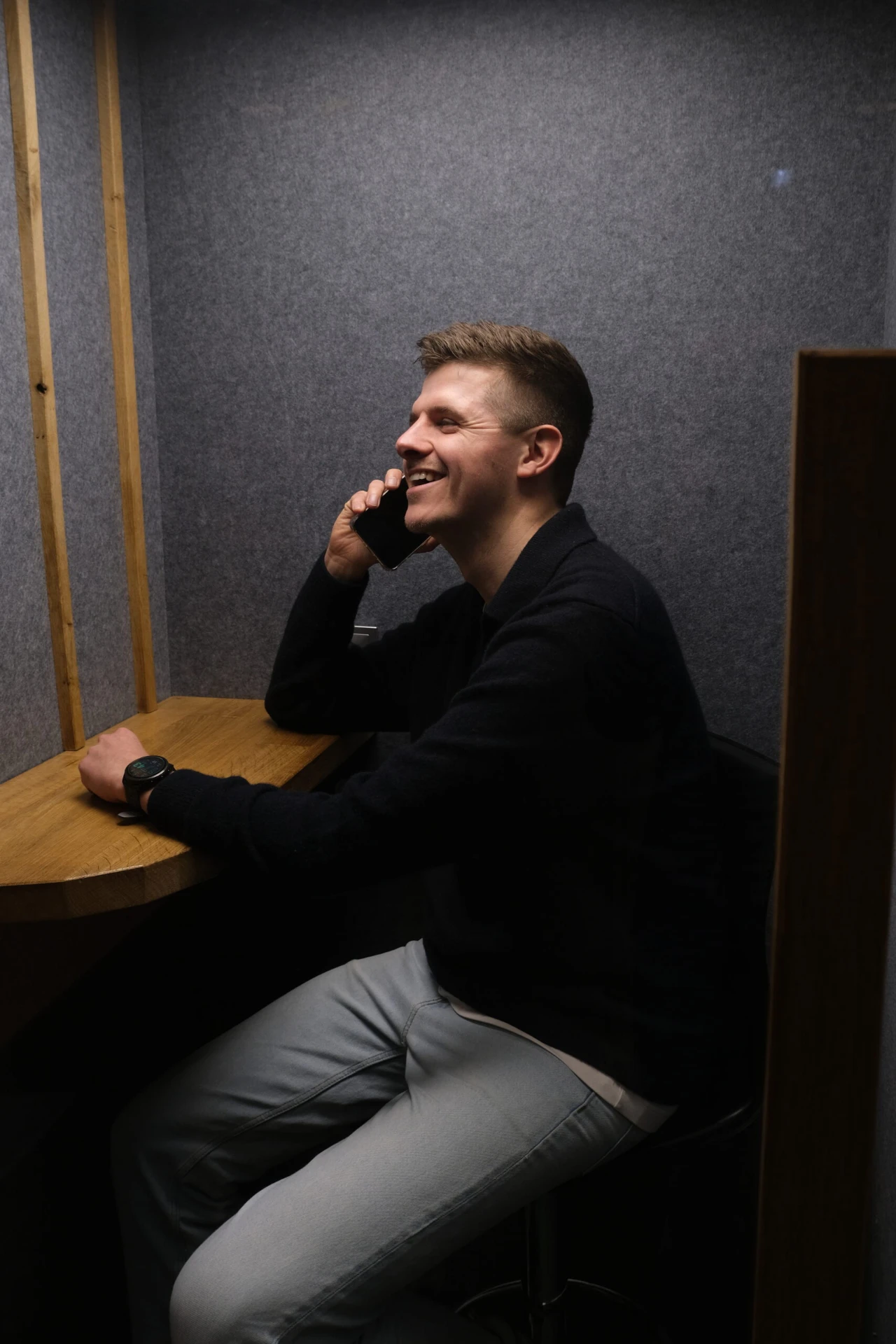 Lachende man belt met zijn smartphone in een akoestische belcel aan een houten bureau.